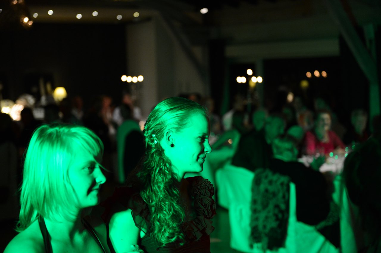 Two women with green lighting on their faces smile at a wedding reception with guests seated in the background.