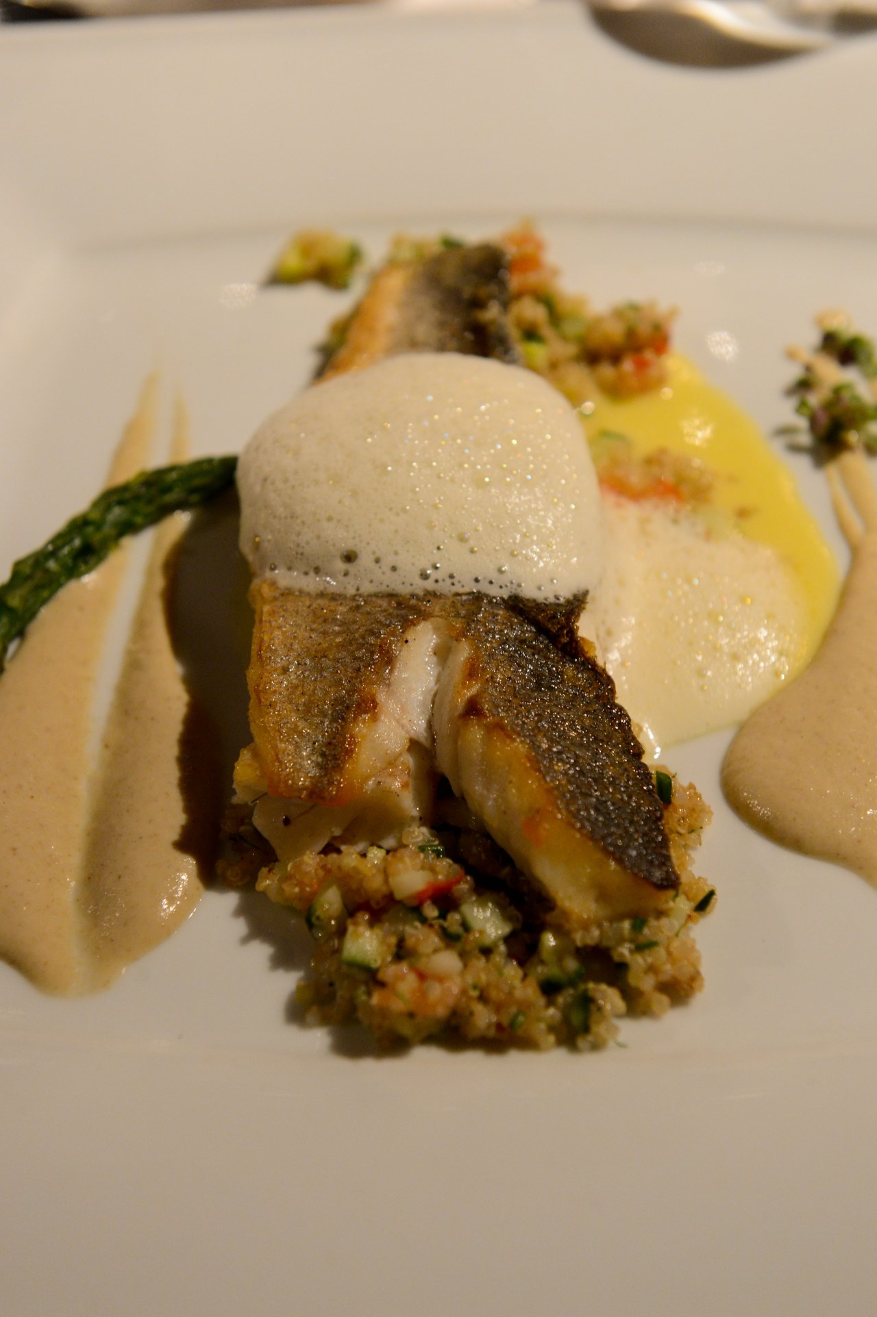 A plated gourmet fish dish with foam, sauce, asparagus, and a side of seasoned grains and vegetables.
