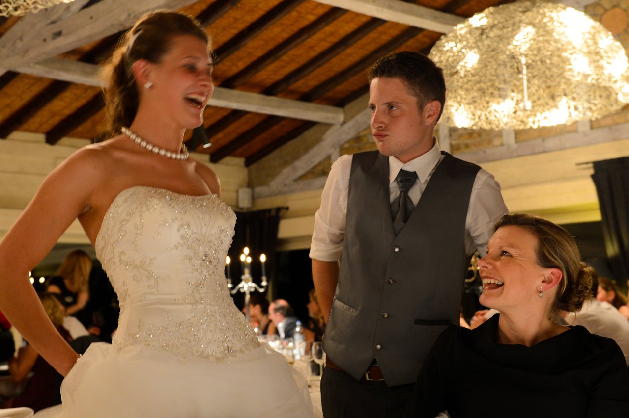 The bride laughs while standing next to a seated guest, as the groom makes a playful expression nearby.