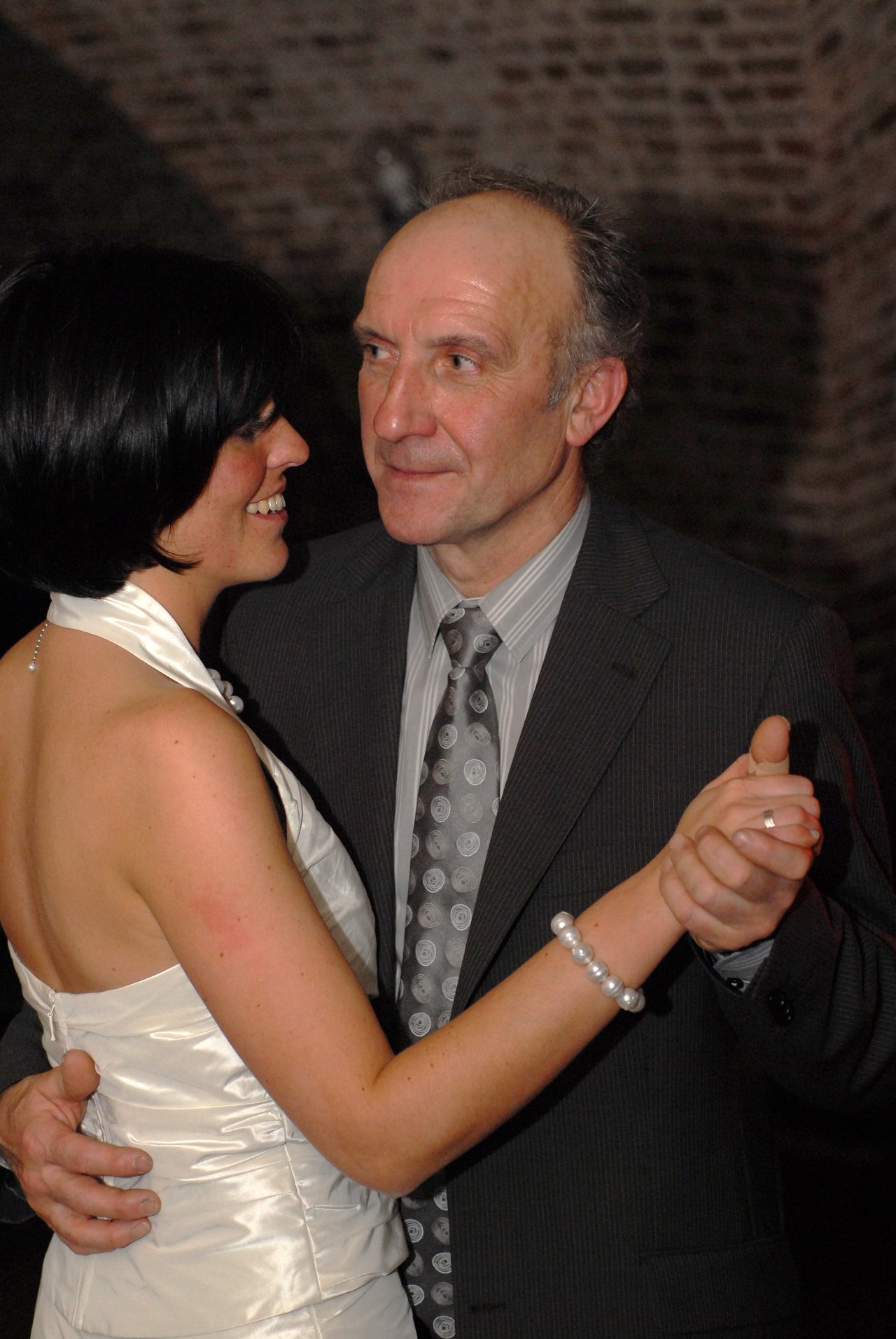 A bride in a white dress dances with an older man in a suit, holding hands and smiling.