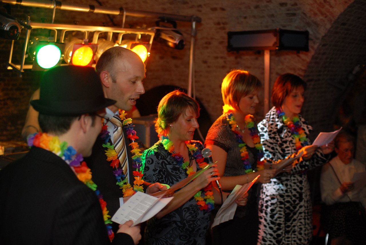 A group of people wearing colorful leis sing together while holding papers at a wedding celebration.