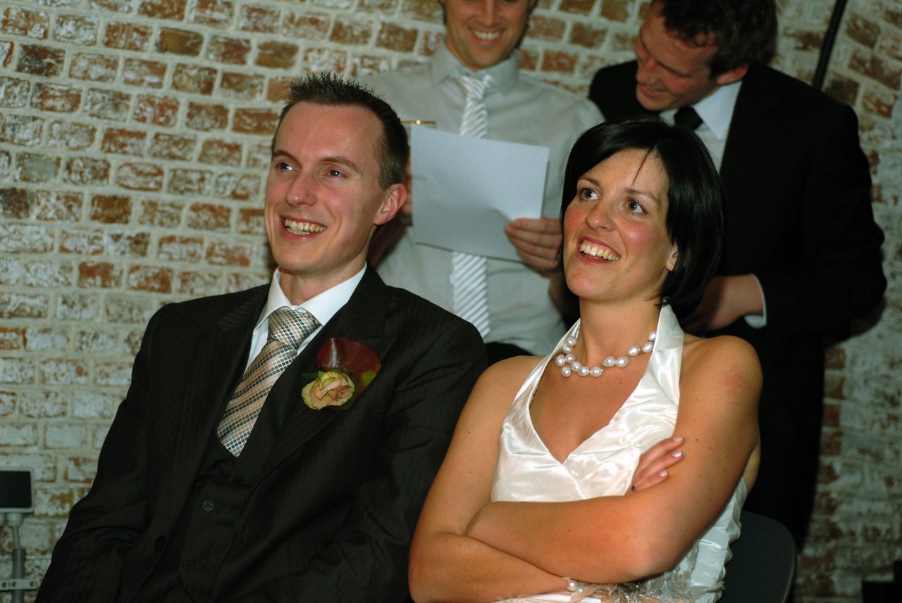 The bride and groom sit together, smiling and laughing, while two men behind them read from a paper.