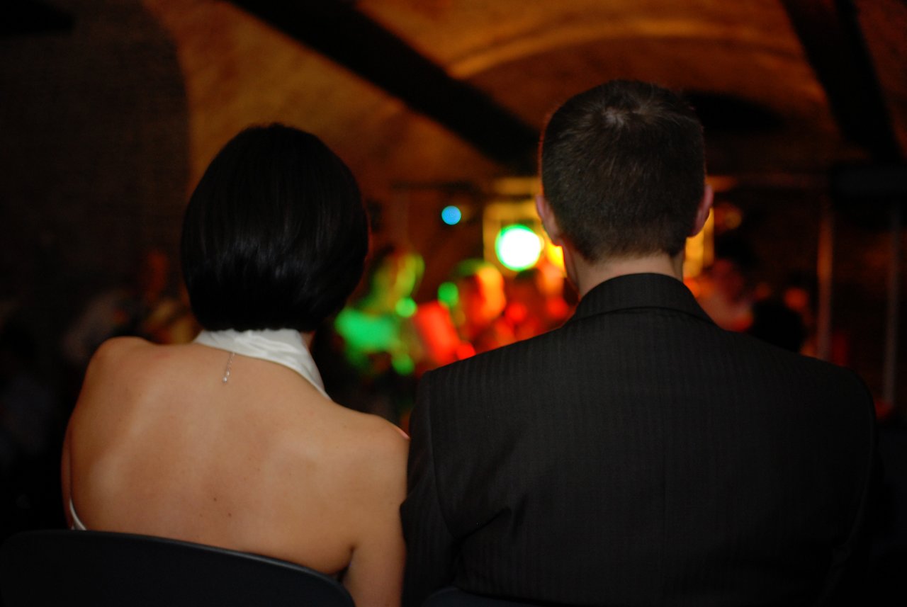 A bride and groom sit together, facing a lively wedding reception with colorful lights and guests in the background.