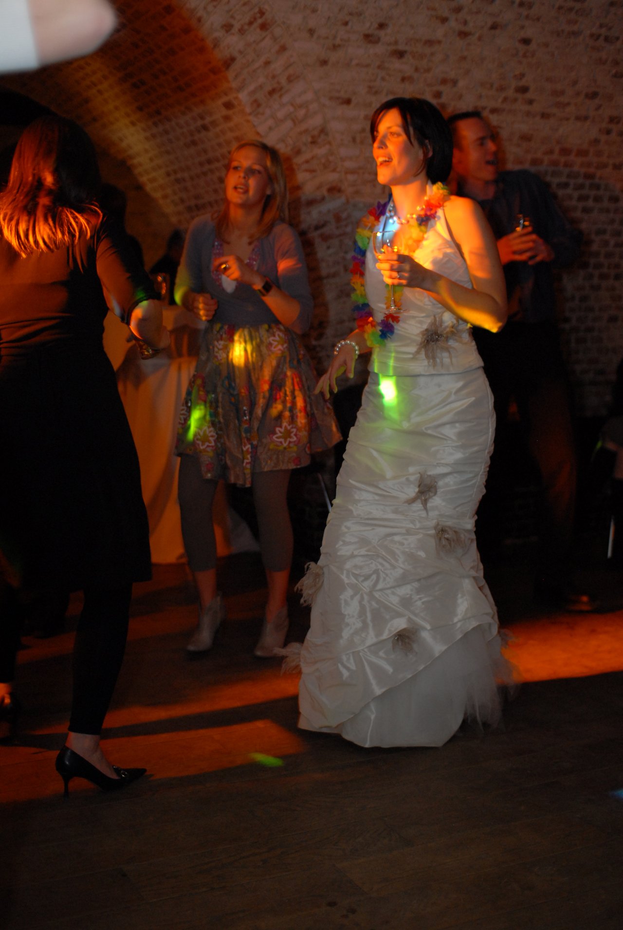 The bride in a white wedding dress dances with guests, holding a drink and wearing a colorful lei.
