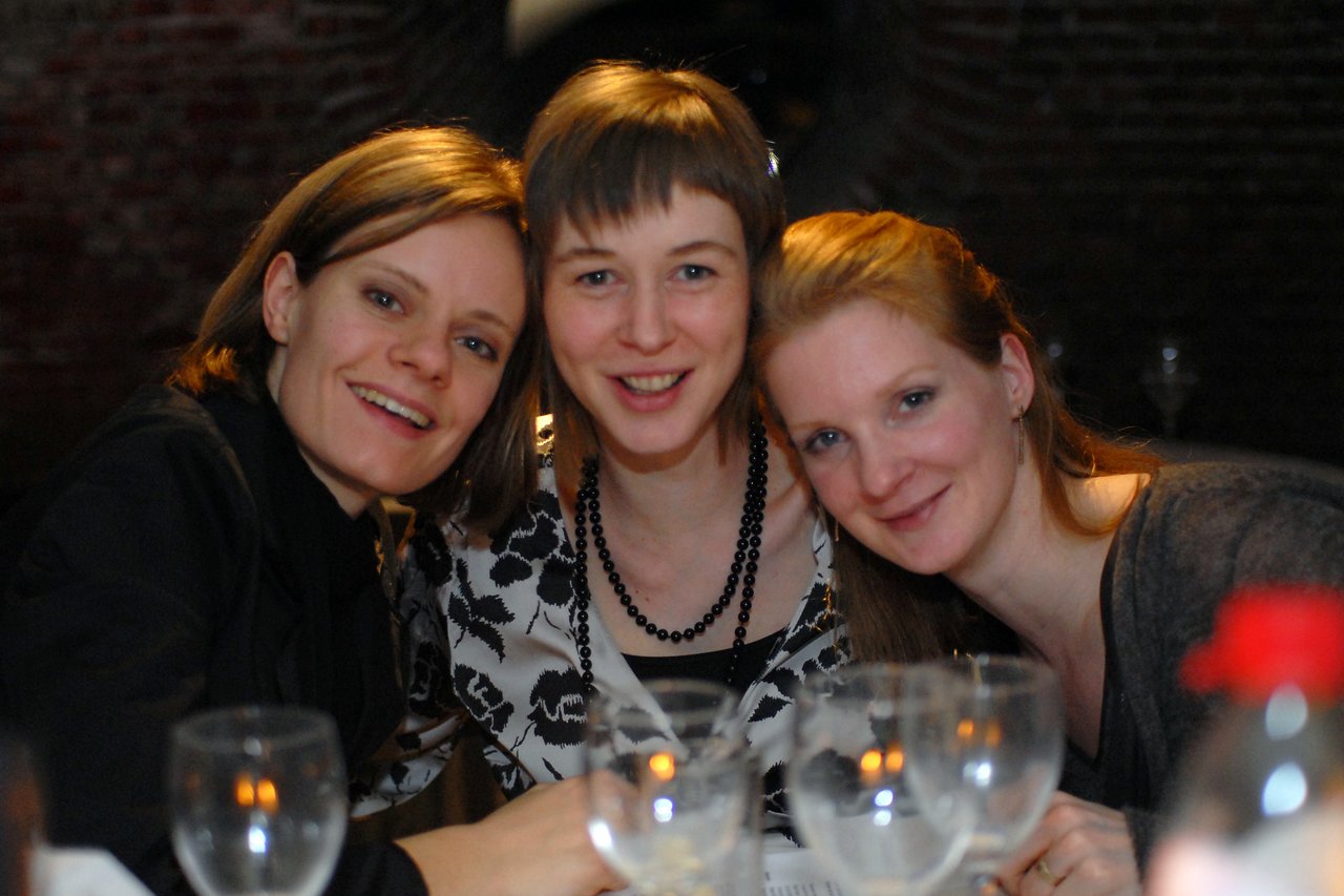 Three smiling women sit closely together at a wedding reception, looking at the camera with drinks on the table.