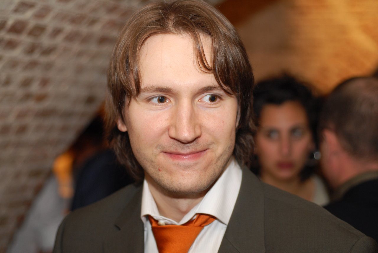 A man in a suit and orange tie smiles while looking to the side at a wedding reception.
