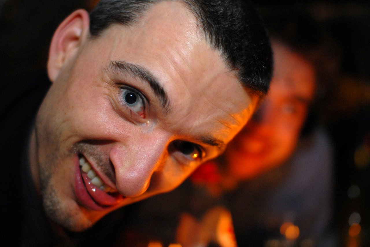 A man leans close to the camera, smiling playfully, with another person slightly blurred in the background.