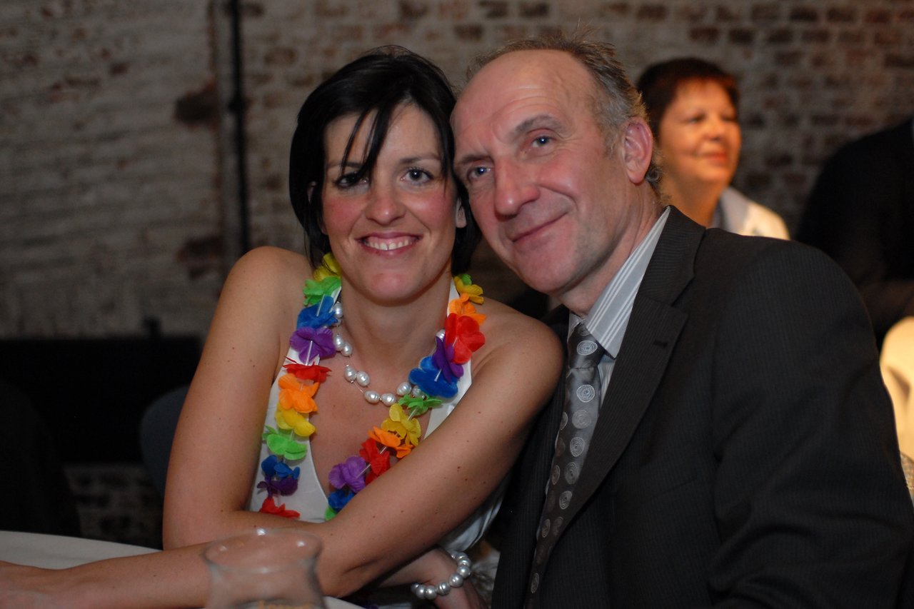 A smiling couple sits closely together at a wedding reception, dressed in formal attire with a colorful flower necklace.