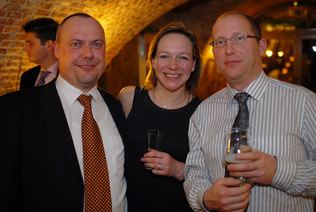 Three people smiling and holding drinks at a wedding reception, dressed in formal attire.