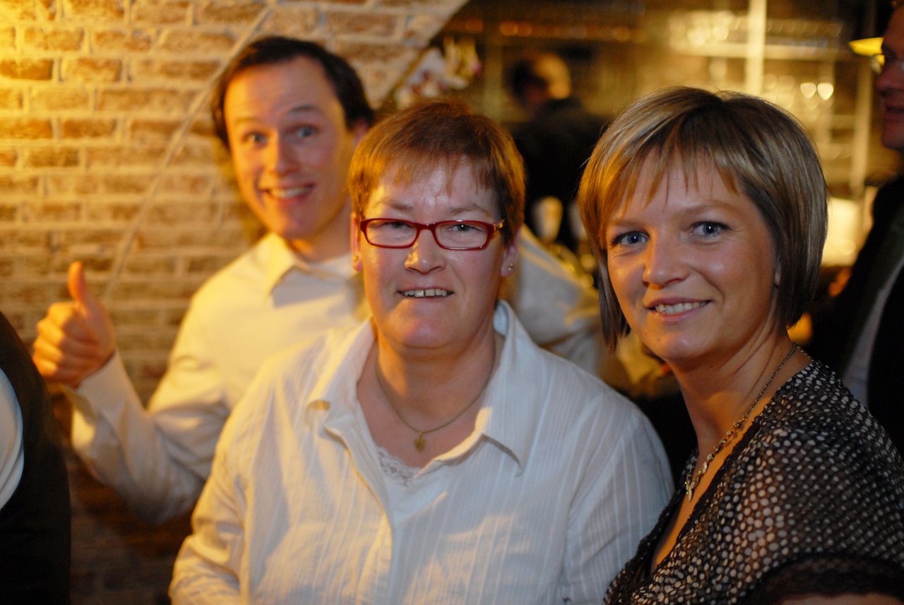 Three people smiling at a wedding reception, with a man in the background giving a thumbs-up.