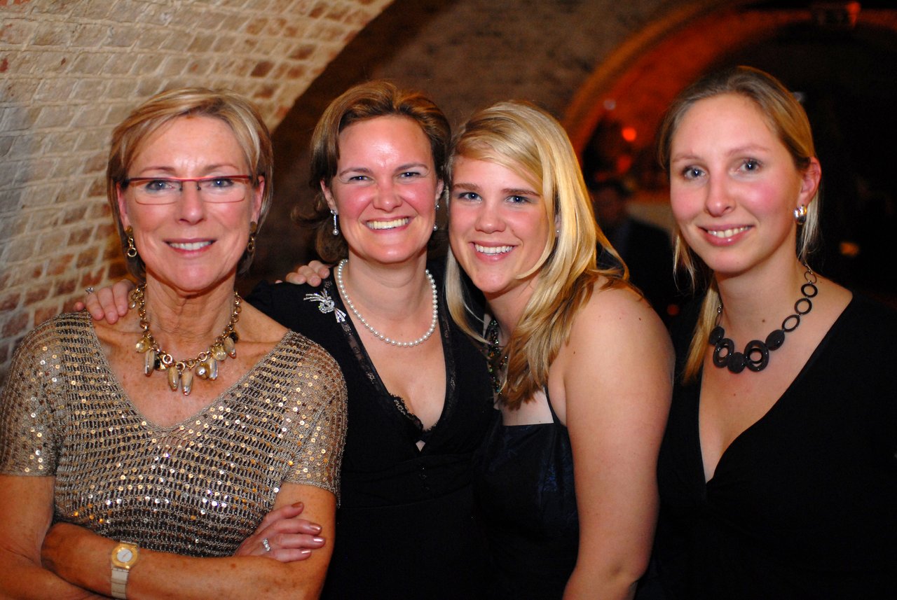 Four women in elegant attire smile and pose together at a wedding celebration in a warmly lit venue.