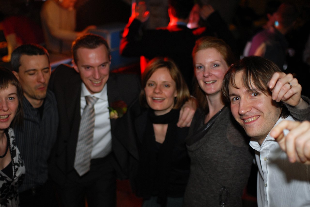 A group of six people smiling and celebrating at a wedding reception, with the groom wearing a suit and boutonniere.