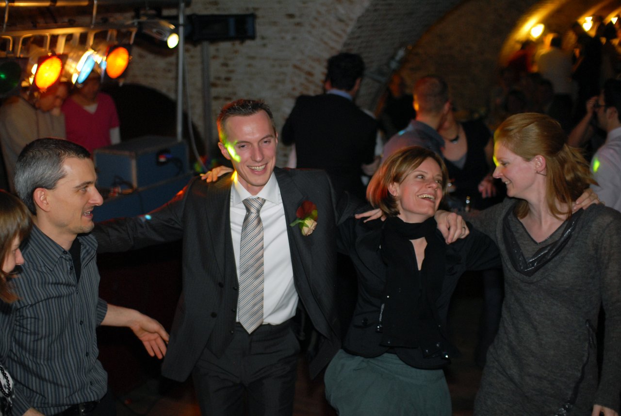 A groom in a suit dances with friends, smiling and celebrating at a wedding reception in a dimly lit venue.