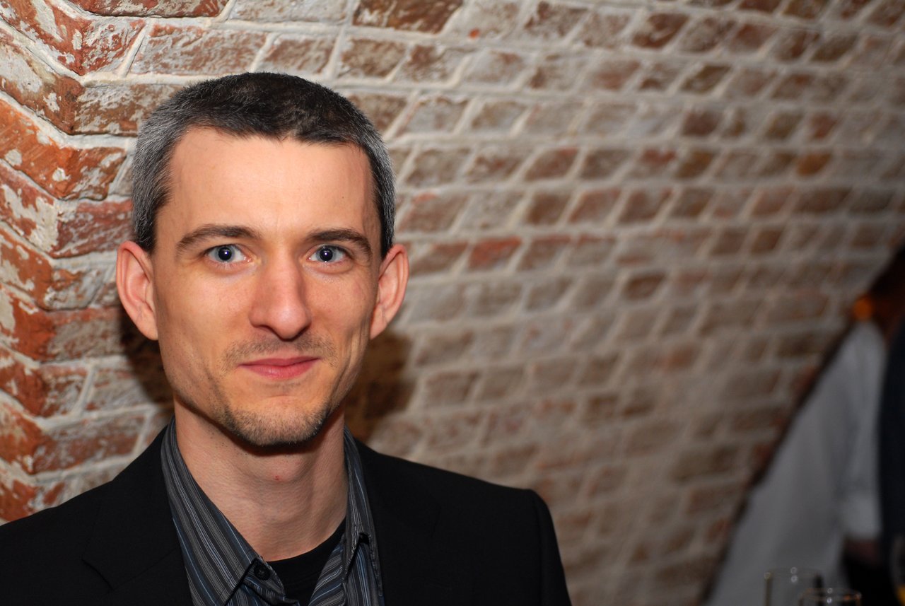 A man in a black suit smiles at the camera during a wedding celebration in a brick-walled venue.