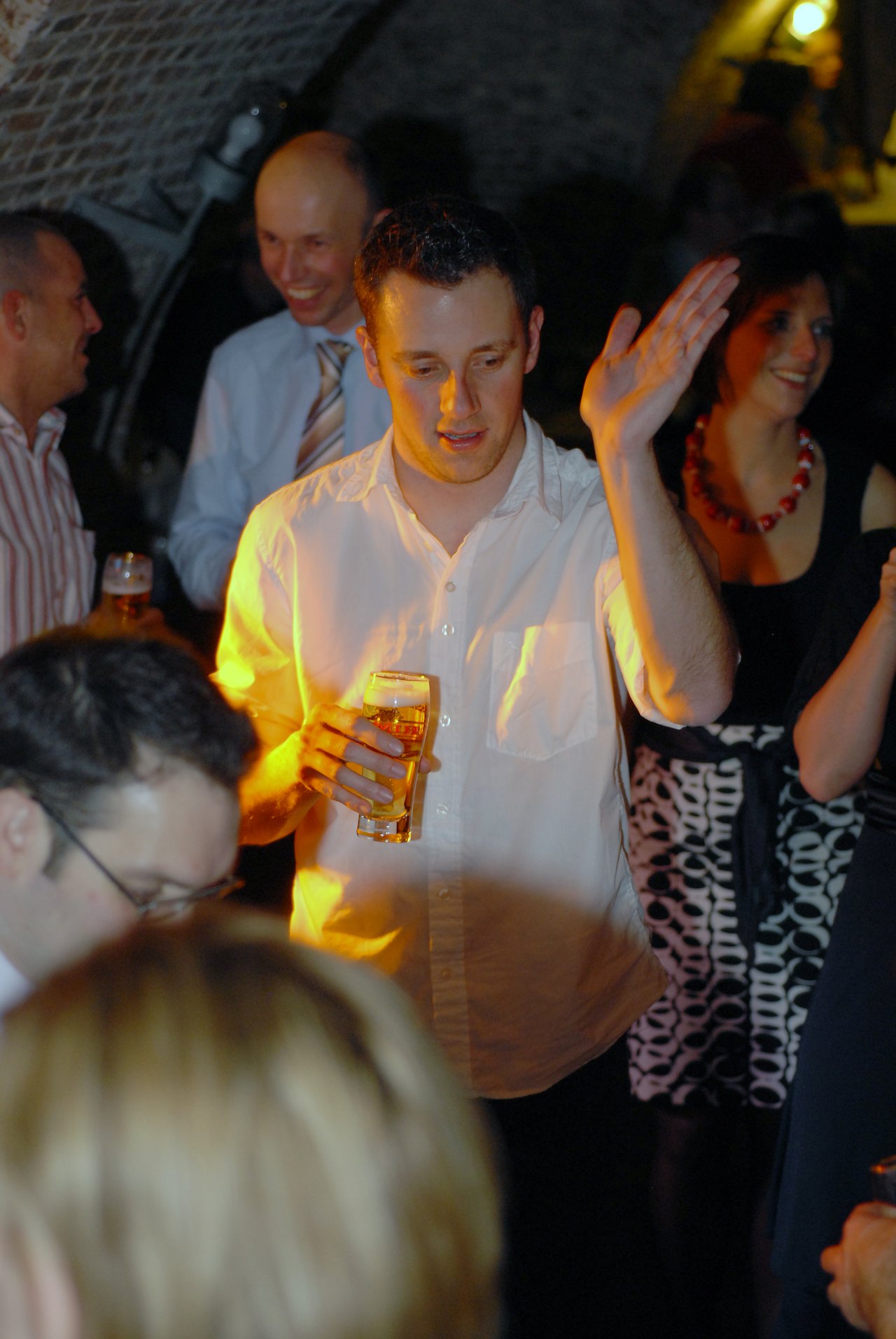 A man in a white shirt holds a beer and gestures while talking at a wedding reception.