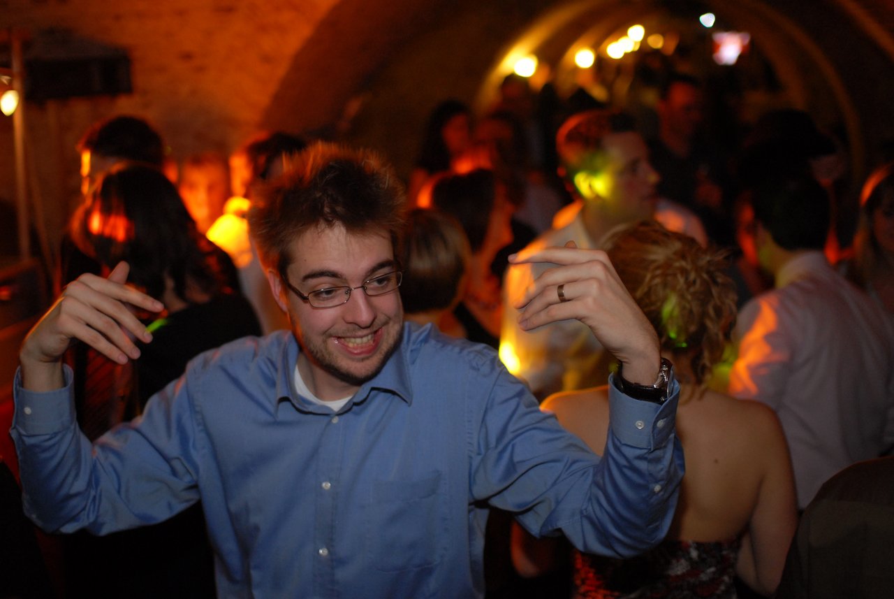 A man in a blue shirt dances and smiles at a wedding reception, surrounded by guests in a dimly lit venue.