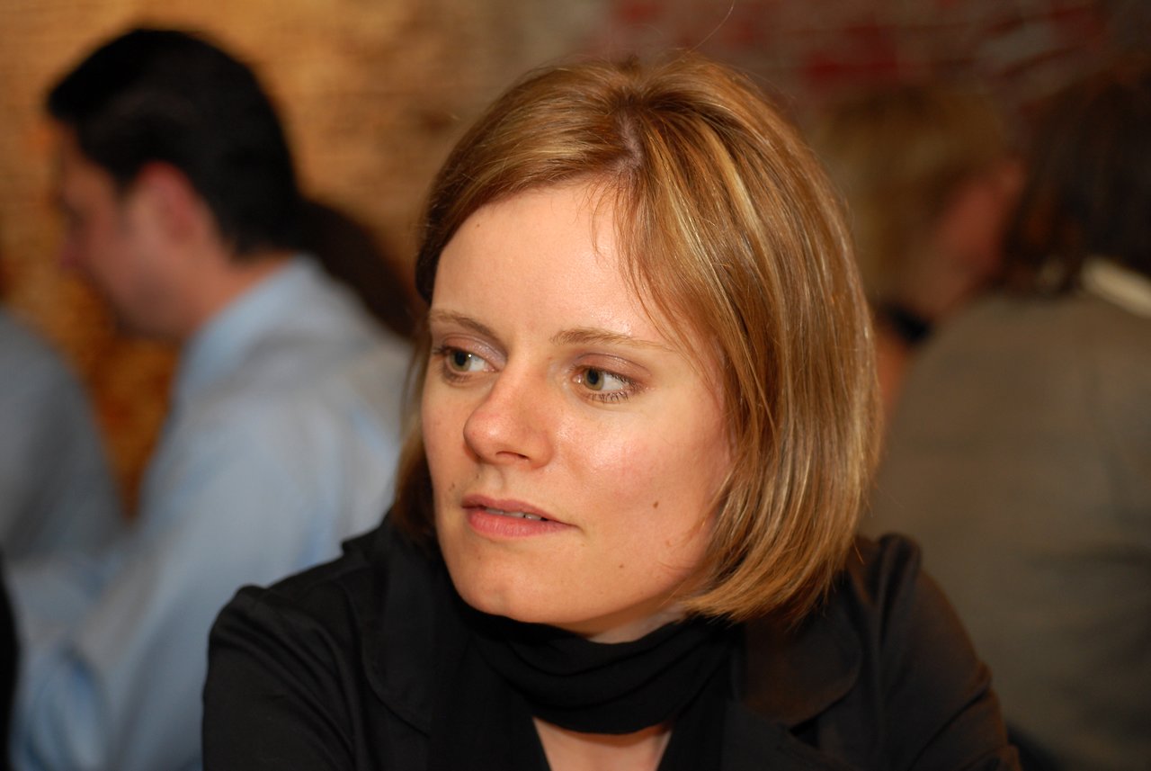 A woman in a black outfit looks to the side at a wedding reception with guests in the background.
