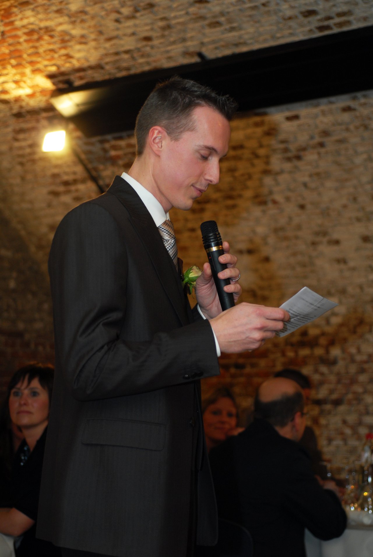 A man in a suit gives a speech at a wedding reception, holding a microphone and a piece of paper.