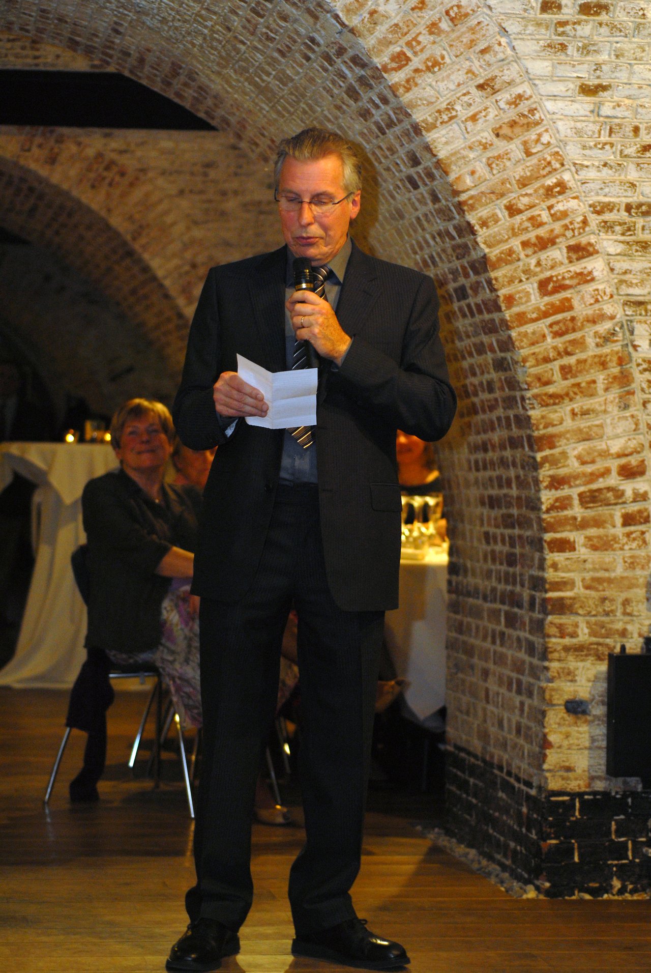 A man in a suit gives a speech at a wedding reception, holding a microphone and a piece of paper.