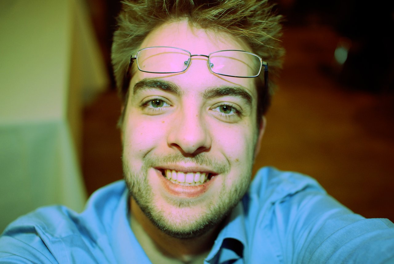 A smiling man in a blue shirt takes a close-up selfie, with glasses resting on his forehead.