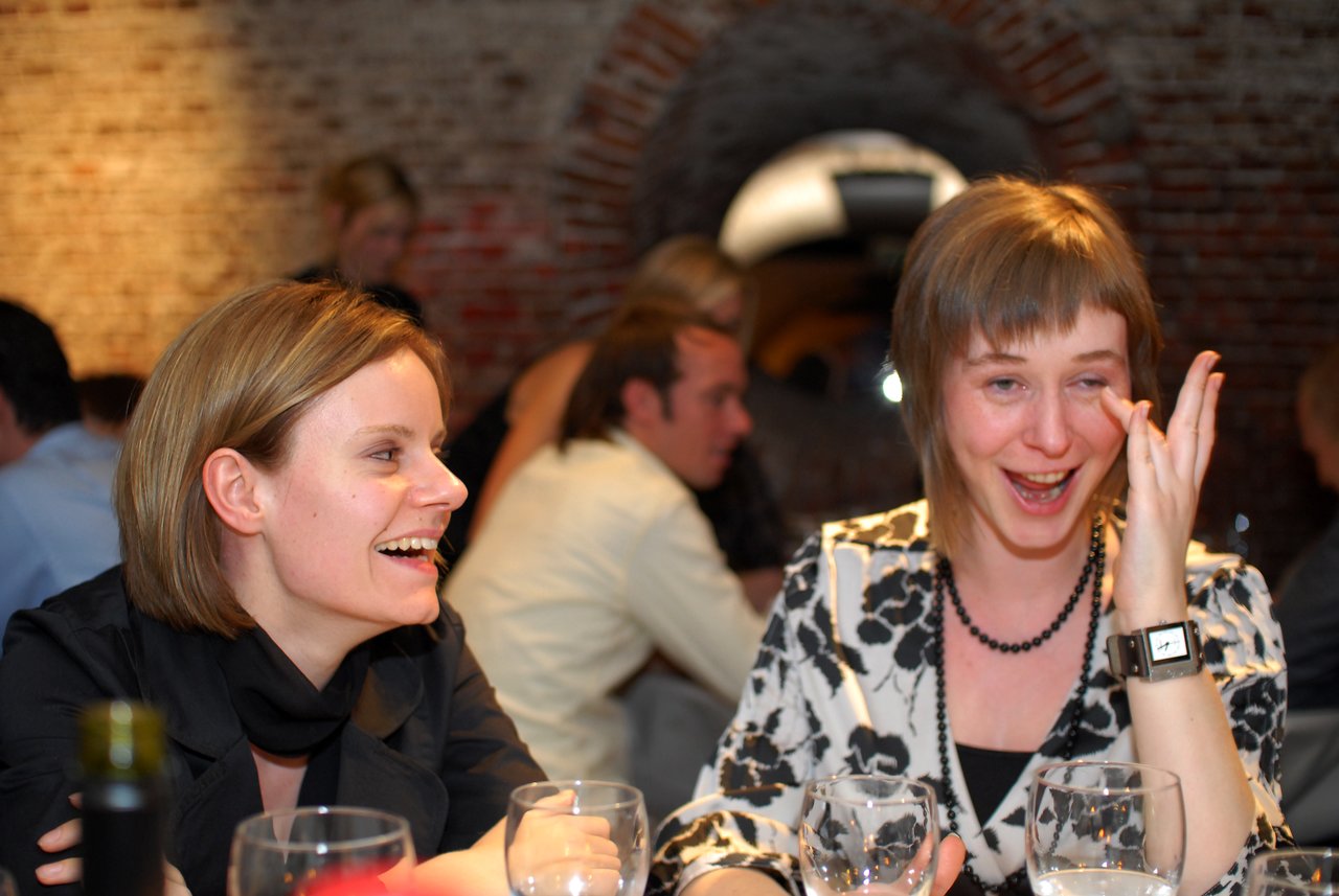 Two women sitting at a table, laughing and smiling, one wiping her eye.
