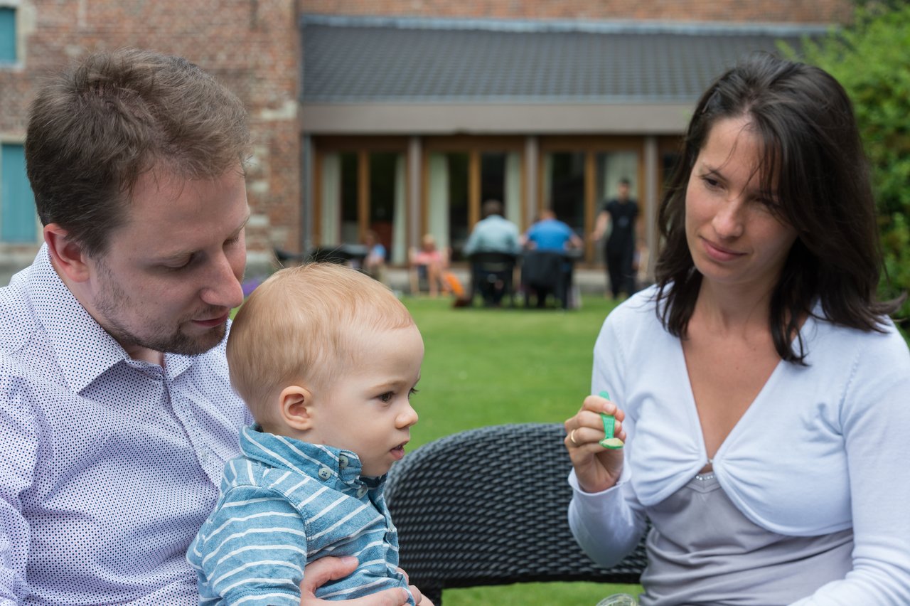 A man holds a young child while a woman nearby holds a small green spoon.