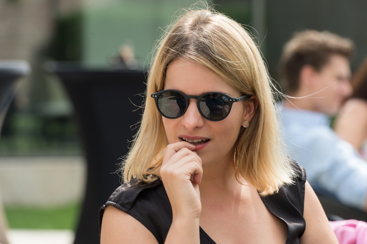 A woman wearing sunglasses and a black dress sits outdoors, touching her chin while looking in a direction.
