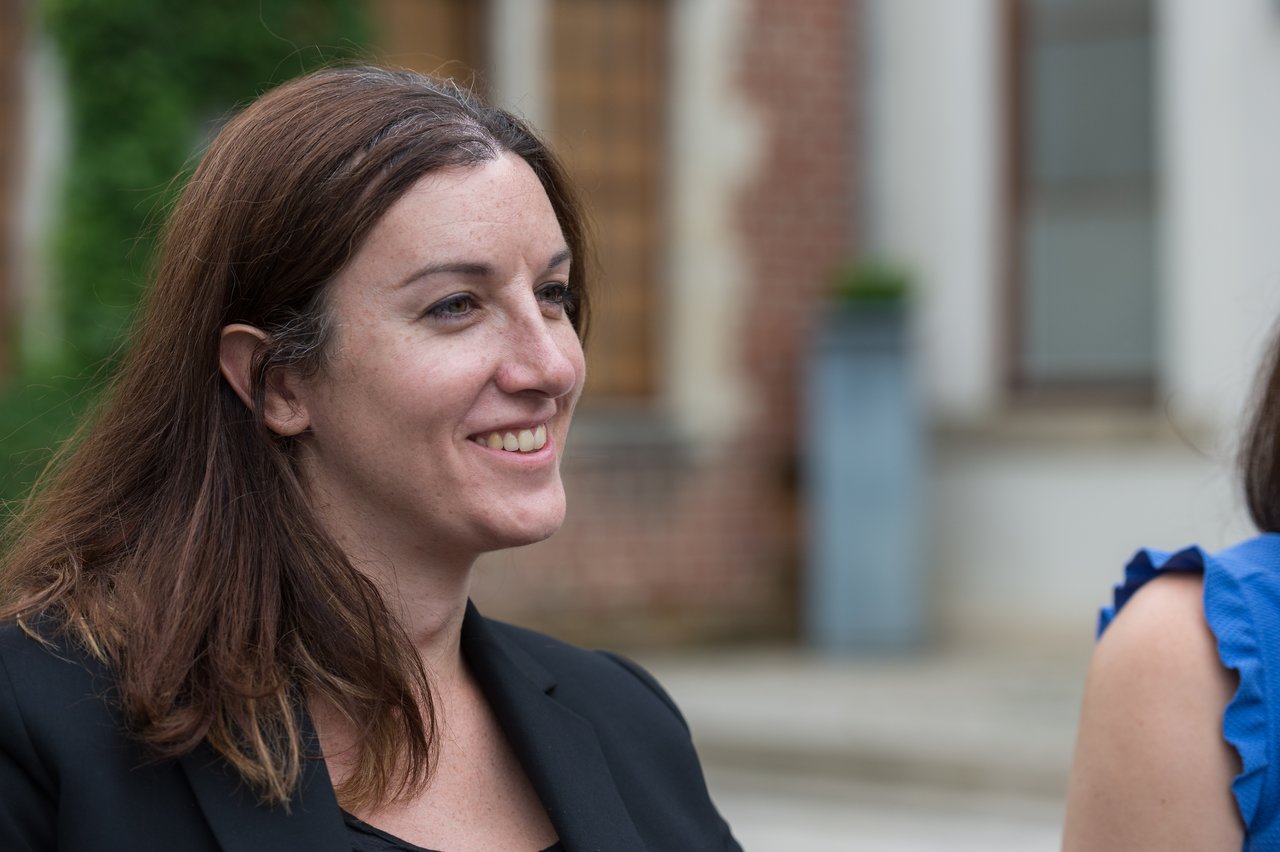 A woman in a black blazer smiles while talking to someone at a wedding.