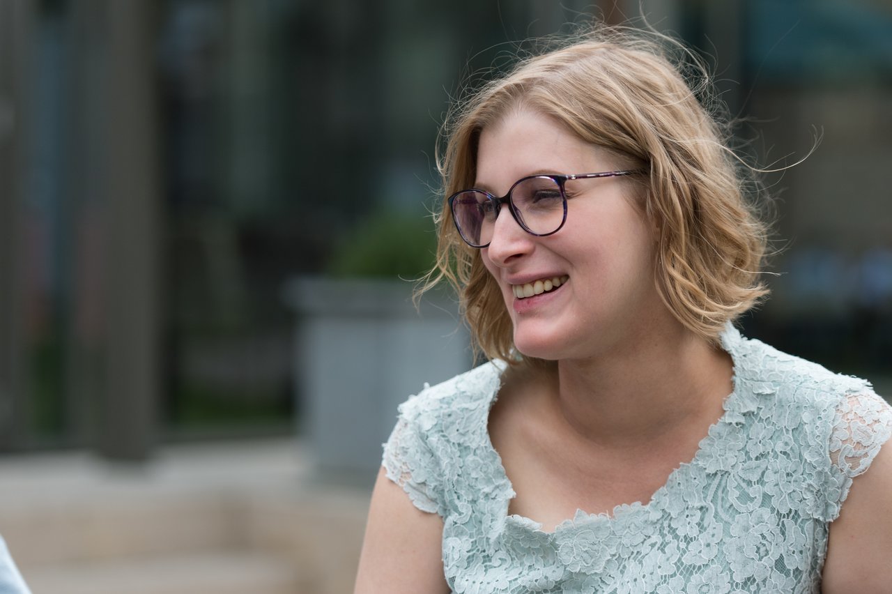 A woman in glasses and a lace dress smiles while looking at someone off-camera.