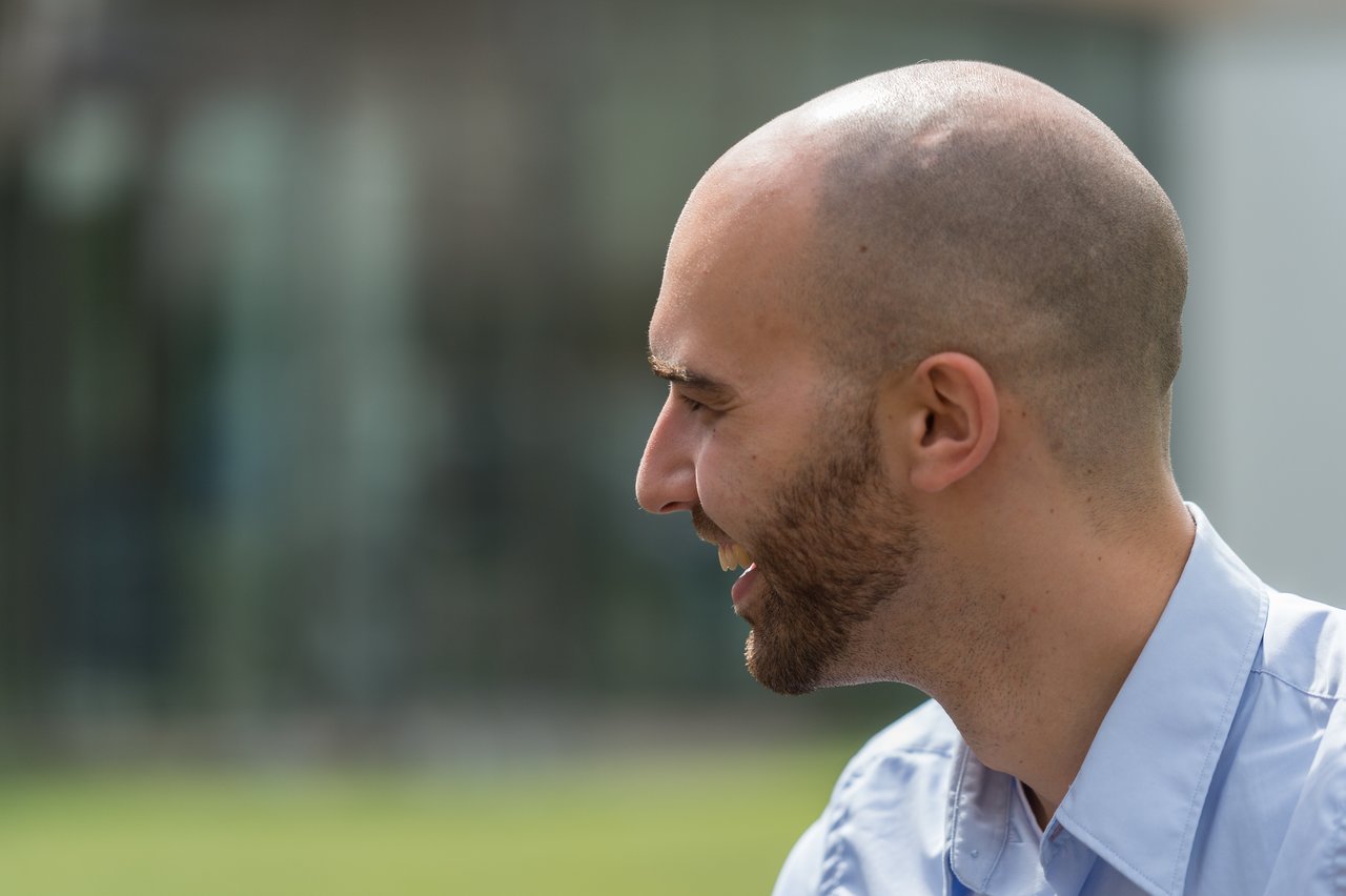 A man in a light blue shirt smiles while looking to the side at an outdoor wedding event.