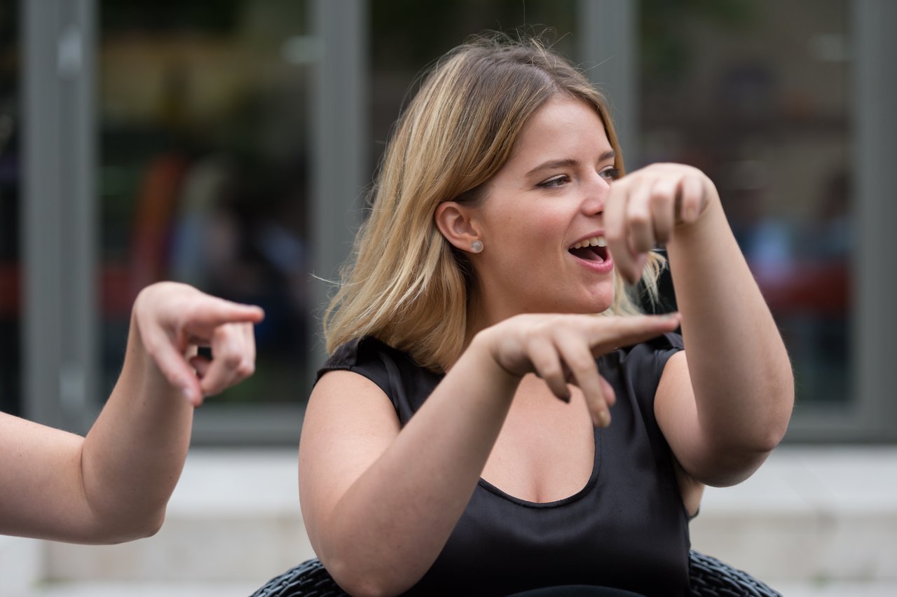 A woman in a black dress is smiling and using hand gestures while communicating with another person.