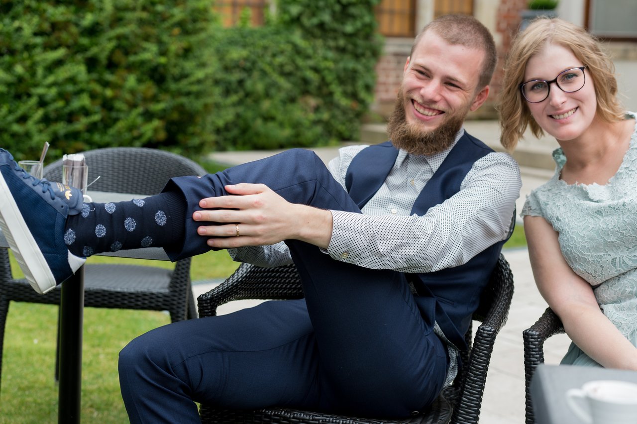 A smiling man in formal attire playfully shows off his patterned socks while sitting next to a smiling woman.