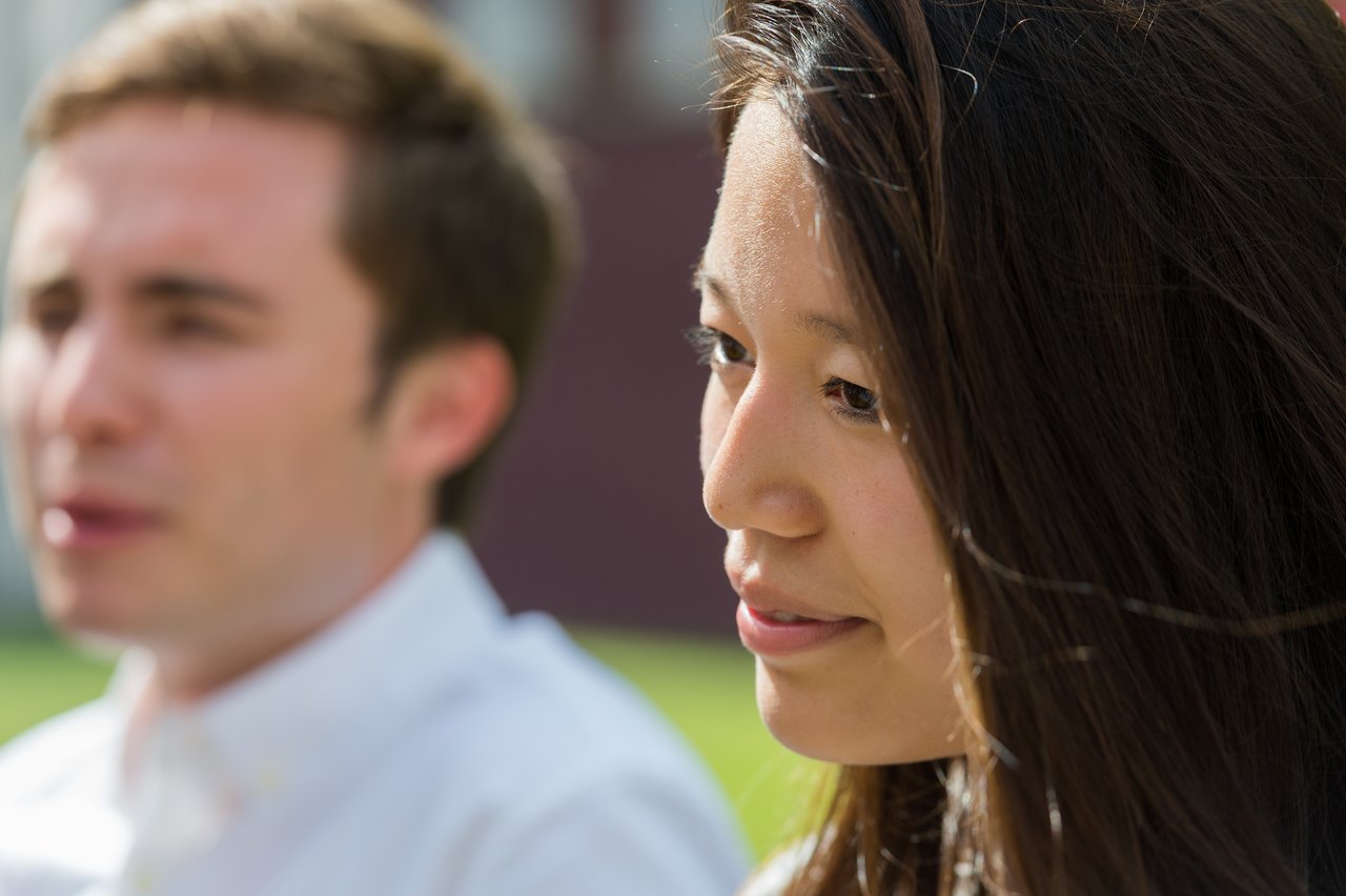A woman looks to the side and smiles, while a man in the background appears to be speaking.