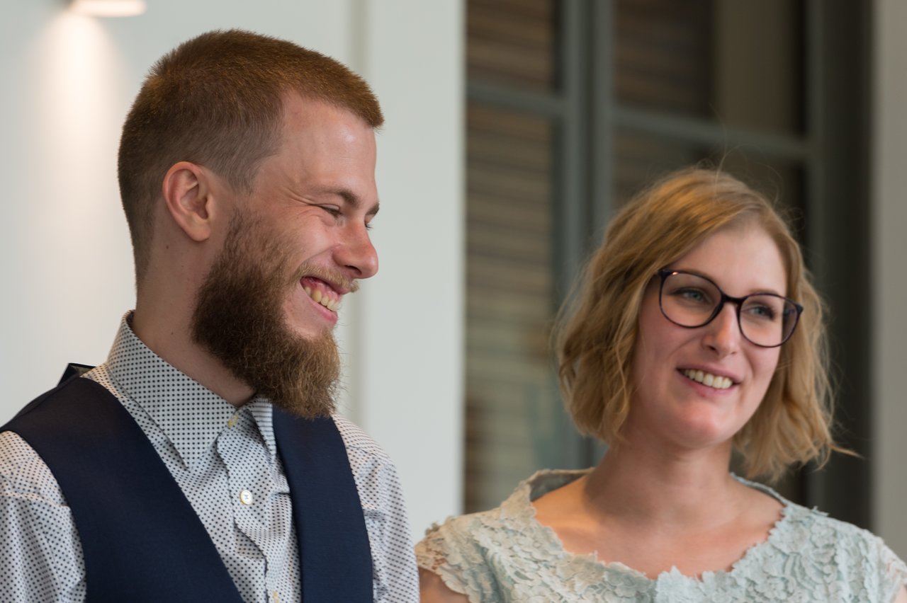 A smiling couple stands together indoors, dressed in formal attire, looking happy on their wedding day.