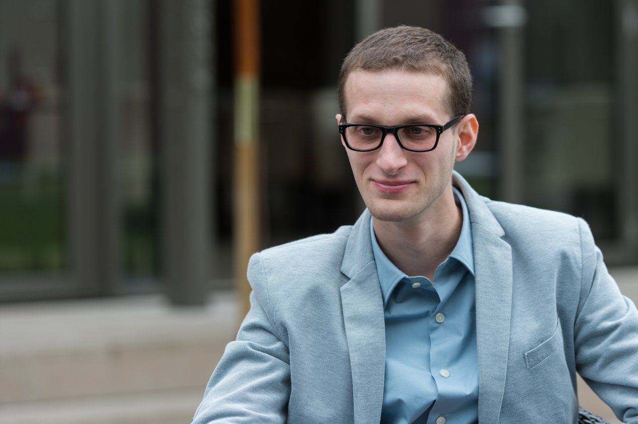 A man in a light blue suit jacket and glasses sits outside, looking to the side with a slight smile.