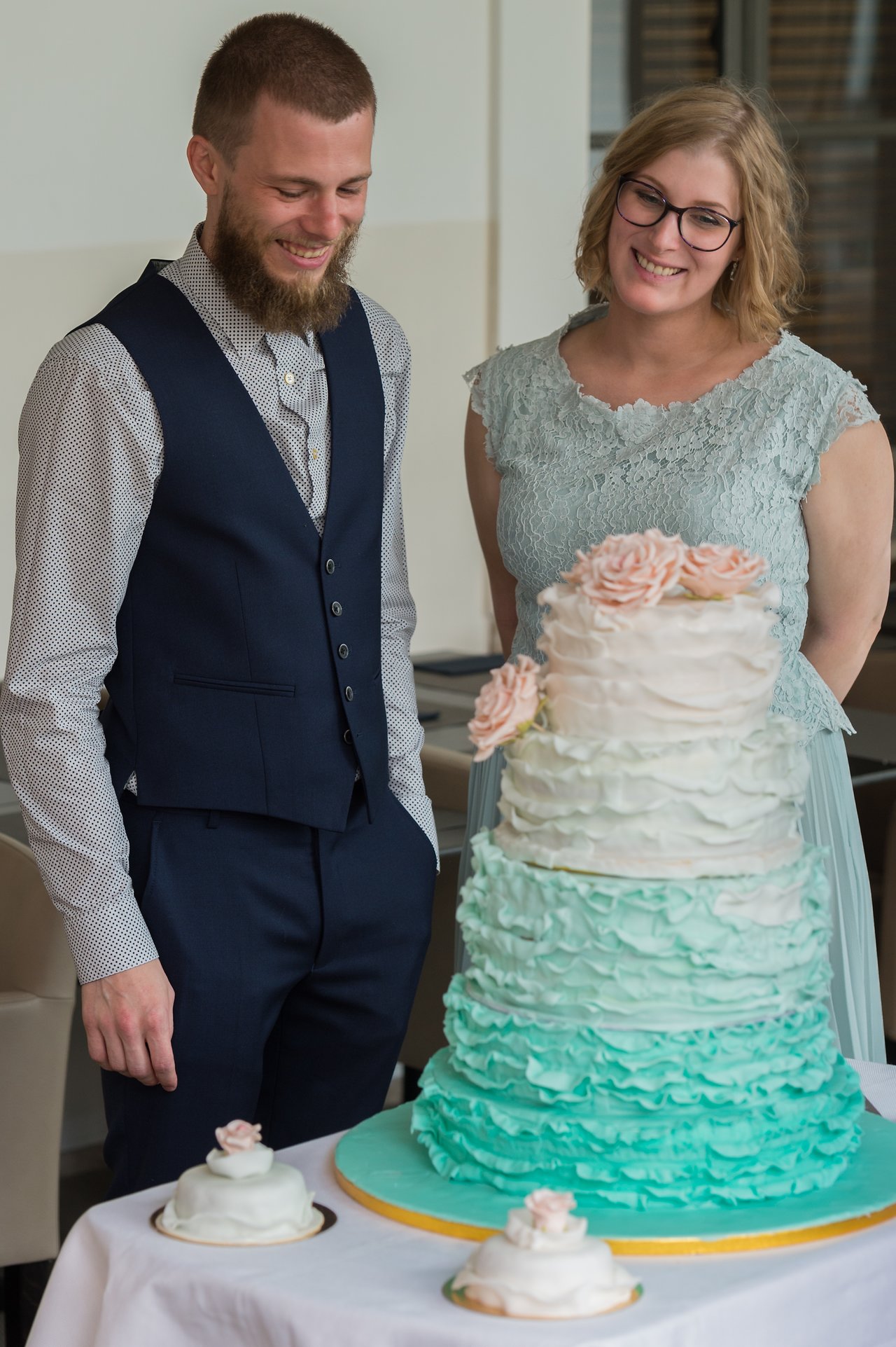 A newlywed couple smiles while looking at their tiered wedding cake decorated with ruffled icing and sugar flowers.