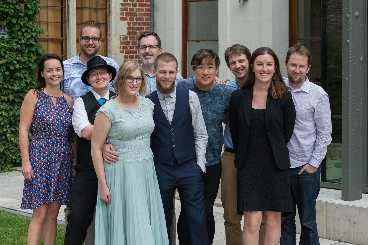 A newlywed couple stands together, smiling, surrounded by friends posing for a group photo at their wedding.