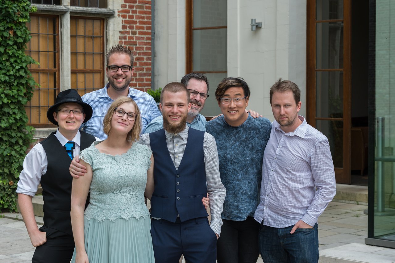 A newlywed couple poses with friends, all smiling and standing together outside a building.