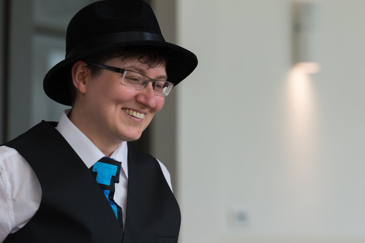 A smiling person wearing a black hat, glasses, and a vest with a blue tie at a wedding.