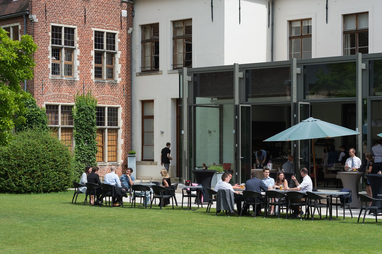 Guests sit at outdoor tables, talking and enjoying drinks at a wedding reception near a modern glass building.