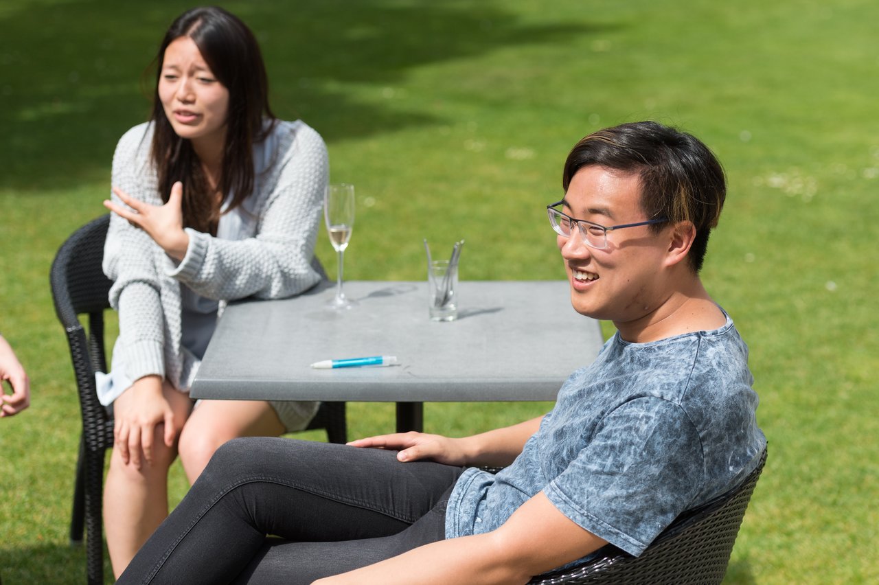 Two people sit at an outdoor table, talking and smiling, with drinks and a pen on the table.