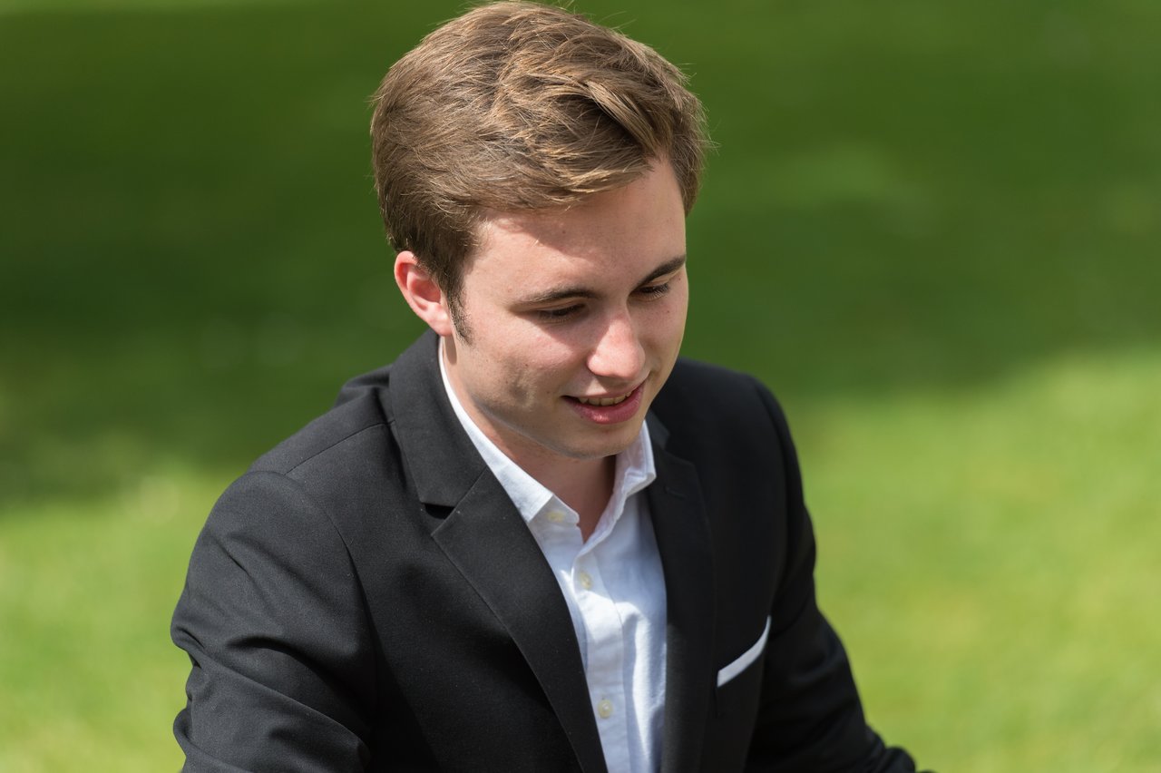 A man in a black suit and white shirt smiles while looking down at something outdoors.