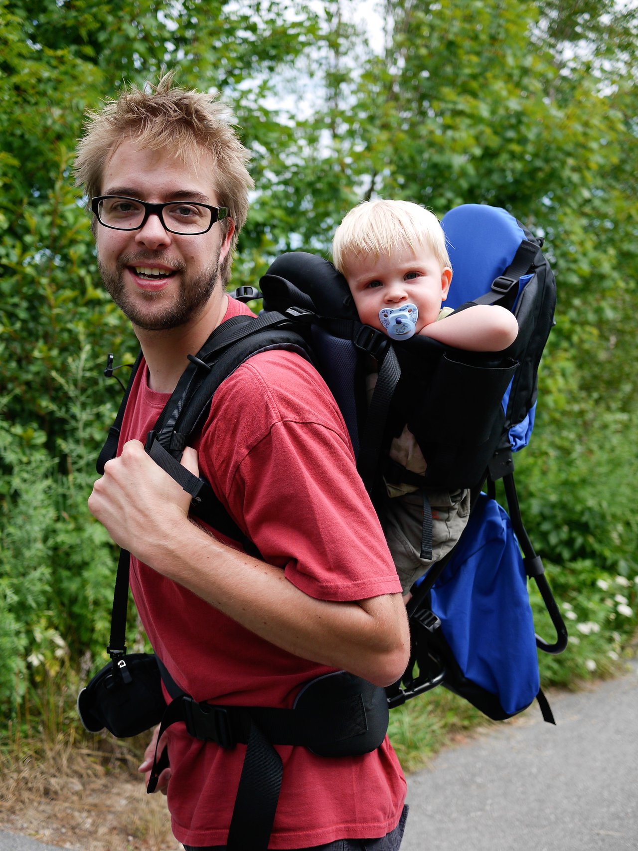 A man wearing a red shirt carries a baby in a backpack carrier while walking outdoors on a paved path.