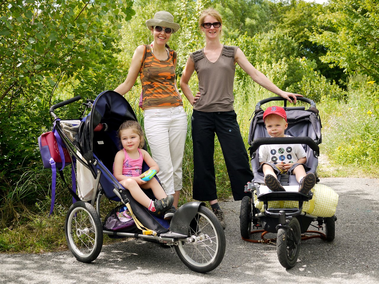 Two women stand on a path with two young children sitting in strollers, surrounded by greenery.