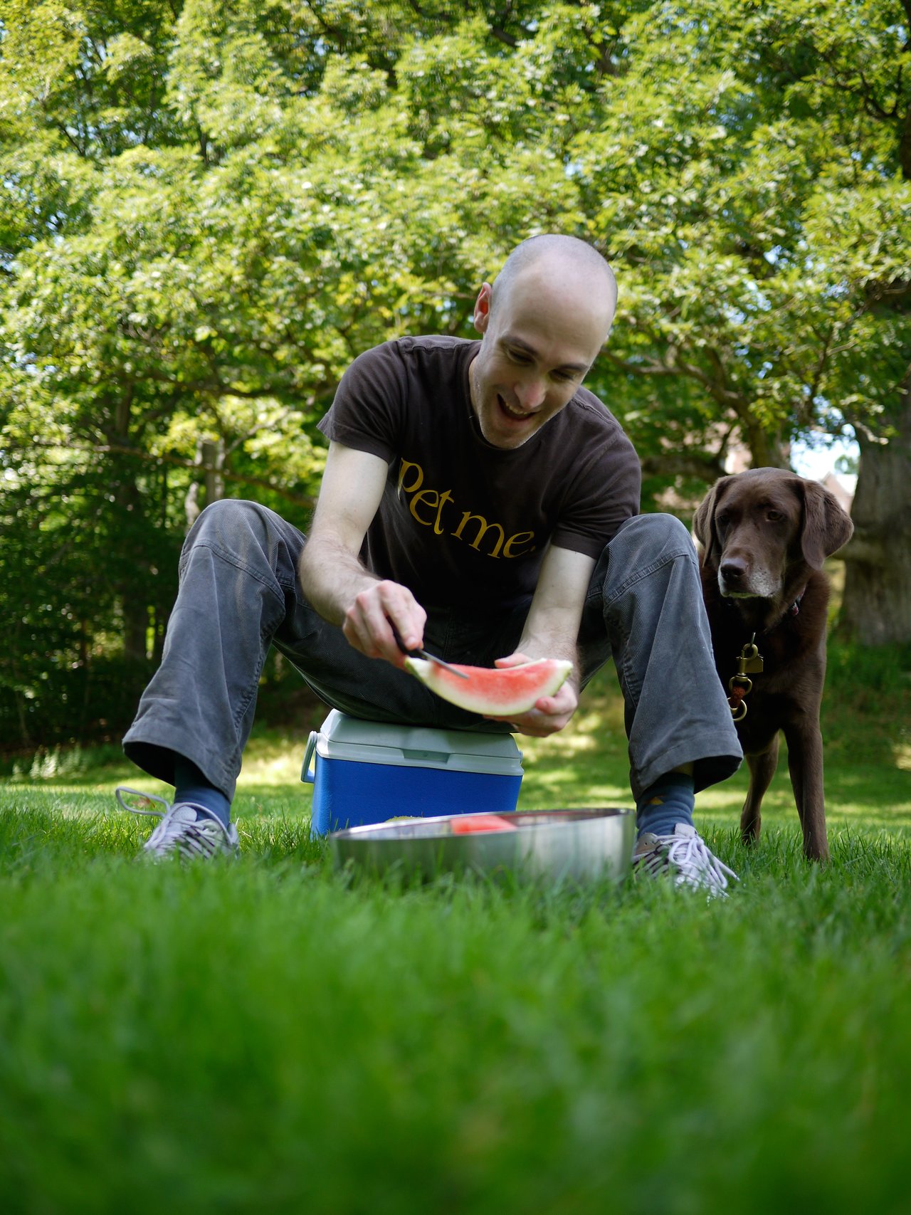 A man sitting on a cooler holds a slice of watermelon while a brown dog stands nearby on the grass.