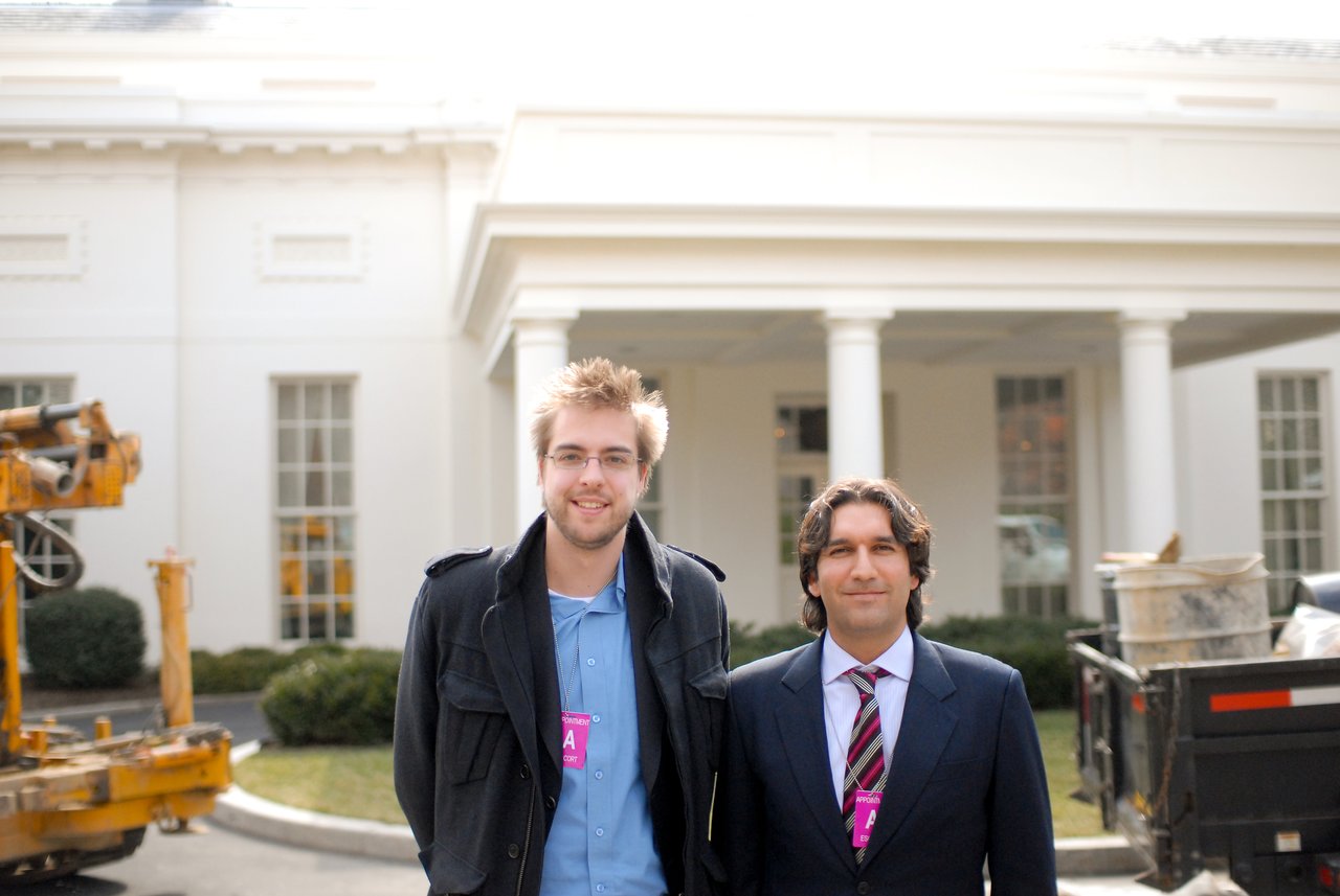 Two men wearing visitor badges stand in front of the White House, smiling at the camera.