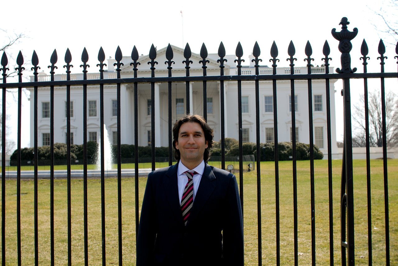 A man in a suit stands in front of a black fence with the White House in the background.