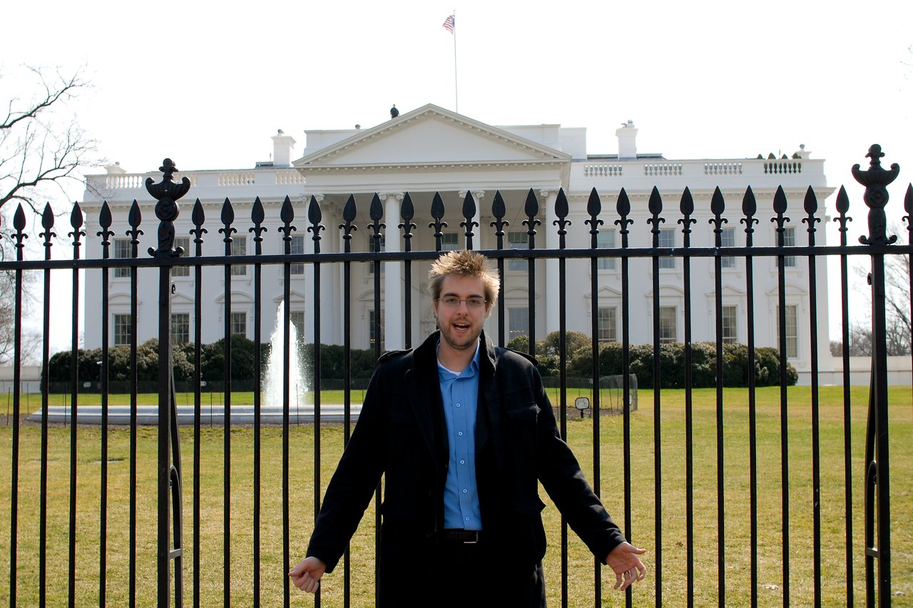 A man stands in front of a black fence with the White House visible behind him.