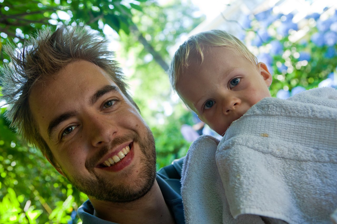 A smiling man holds a young child wrapped in a towel, both looking at the camera in an outdoor setting.