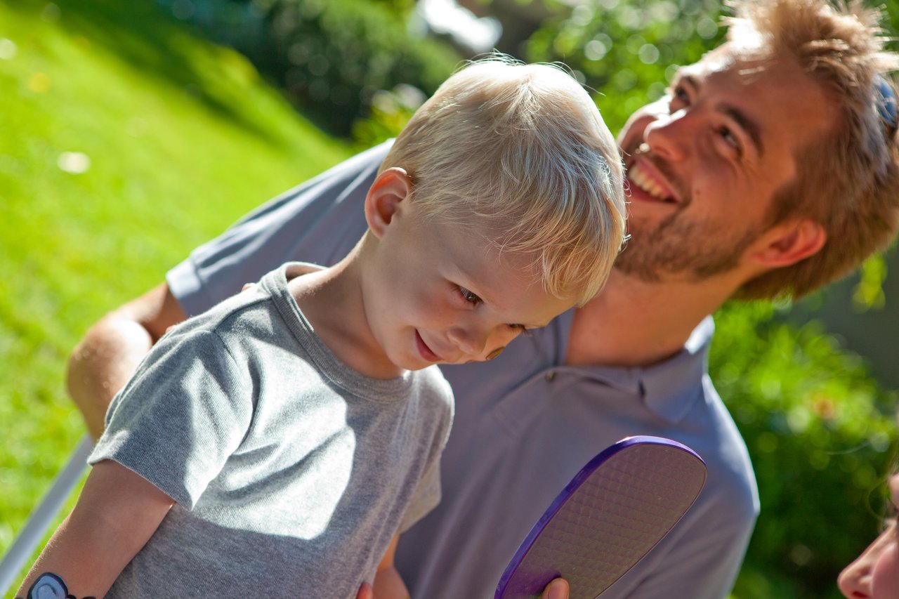 A smiling man stands behind a young boy holding a purple paddle, both appearing happy in an outdoor setting.